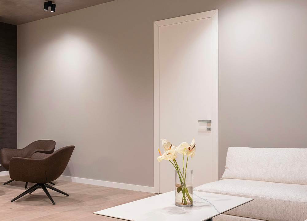 Porta interna bianca a battente inserita in un soggiorno moderno dai toni neutri, con pareti grigio chiaro, arredi minimal e luce soffusa.