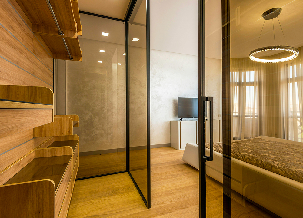 Soluzione moderna con porte interne scorrevoli in vetro fumé per dividere una cabina armadio da una camera da letto open space con parquet chiaro.