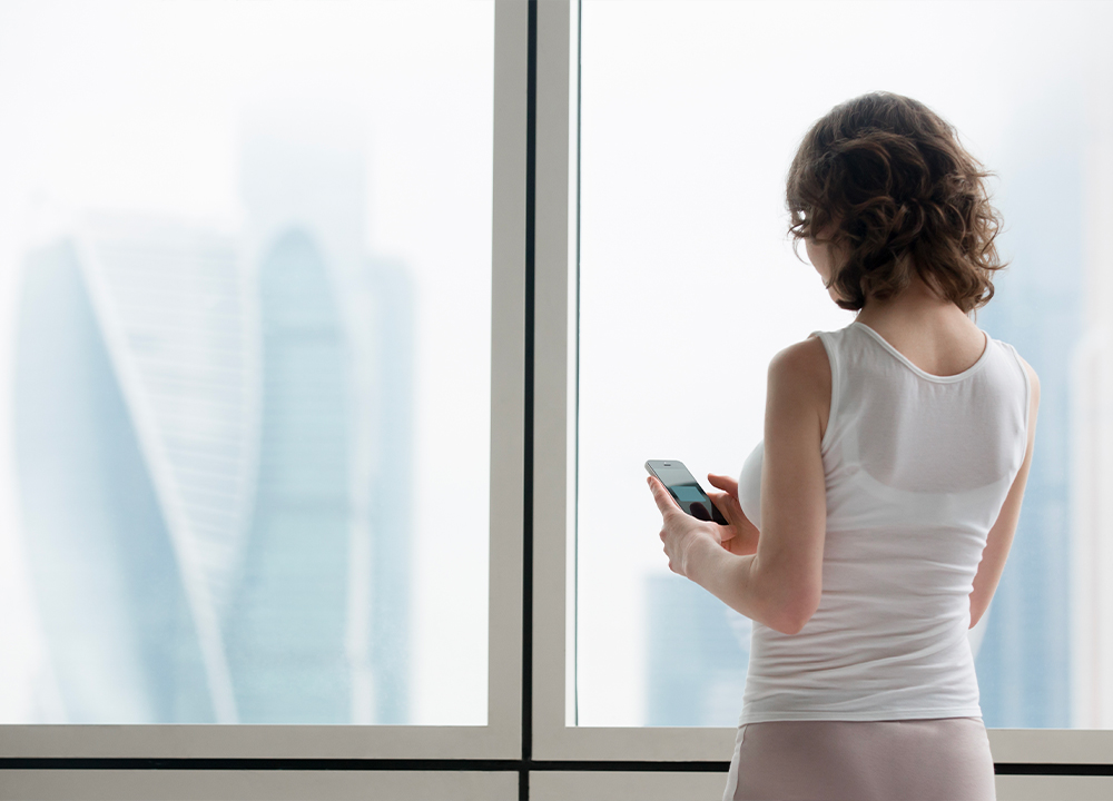Donna che controlla una finestra intelligente con lo smartphone vicino a una vetrata moderna