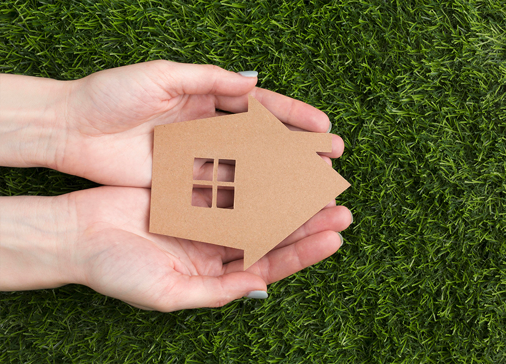 Mani che tengono una piccola casa di cartone su prato verde